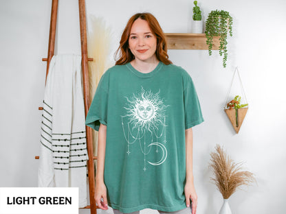 May include: A light green short-sleeve t-shirt featuring a white graphic print of a sun with a human face, surrounded by hanging stars and a crescent moon. The design is centered on the chest of the garment, which has a crew neckline.