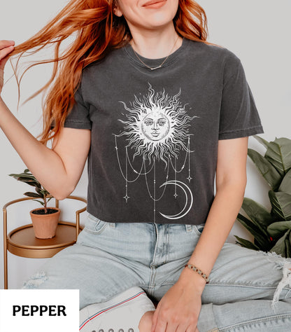 May include: A charcoal gray short-sleeve t-shirt featuring a white graphic print of a sun with a human face, surrounded by radiating rays, hanging beaded chains, and a crescent moon. The shirt has a crew neckline and a relaxed, casual fit.