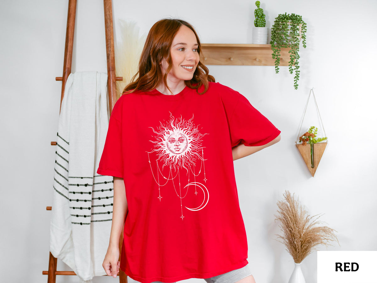 May include: A bright red short-sleeved t-shirt featuring a large white graphic print on the front. The design depicts a stylized sun with a human face surrounded by radiating rays, with dangling chains, small stars, and a crescent moon below it.