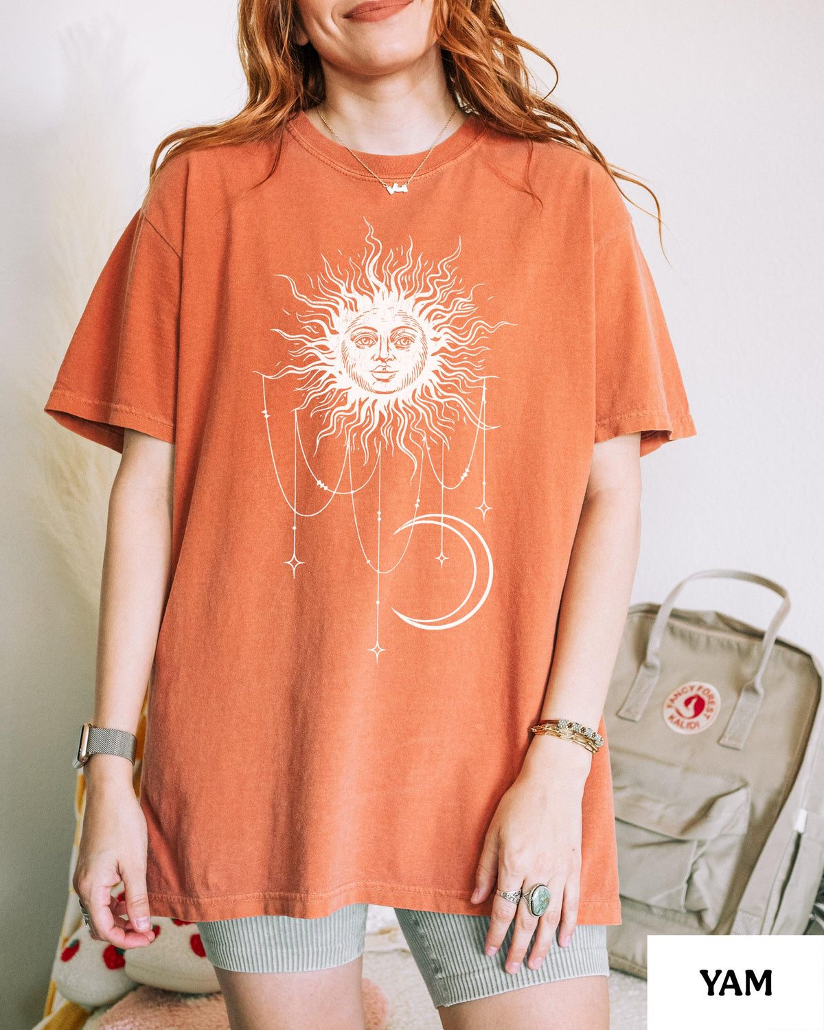 May include: A burnt orange short-sleeve t-shirt featuring a white graphic print of a sun with a face, a crescent moon, and hanging beaded chains with star accents. The shirt has a crew neckline and a relaxed fit, showcasing a celestial design.