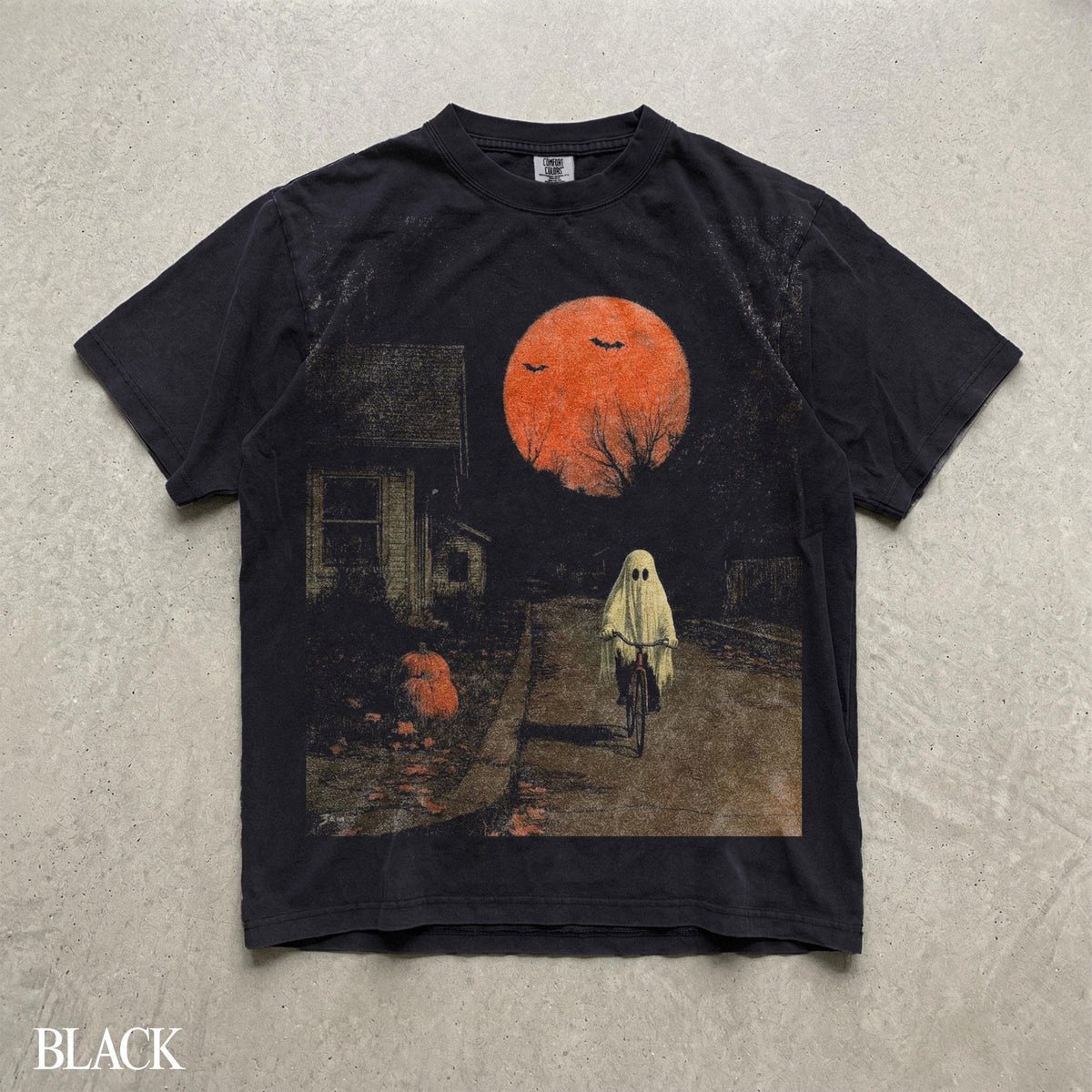 May include: A black short-sleeve t-shirt featuring a graphic print of a ghost riding a bicycle on a road at night. The scene includes a large orange moon, two flying bats, a house on the left, and a pumpkin on the ground near the sidewalk.