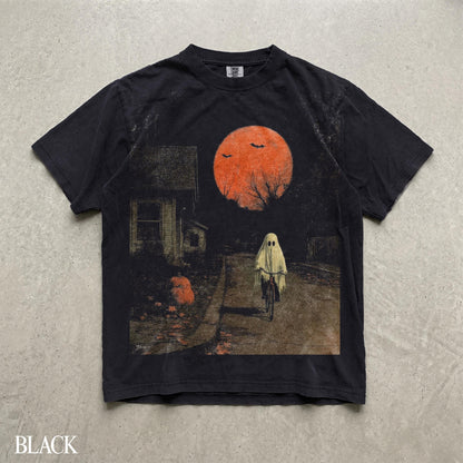 May include: A black short-sleeve t-shirt featuring a graphic print of a ghost riding a bicycle on a road at night. The scene includes a large orange moon, two flying bats, a house on the left, and a pumpkin on the ground near the sidewalk.