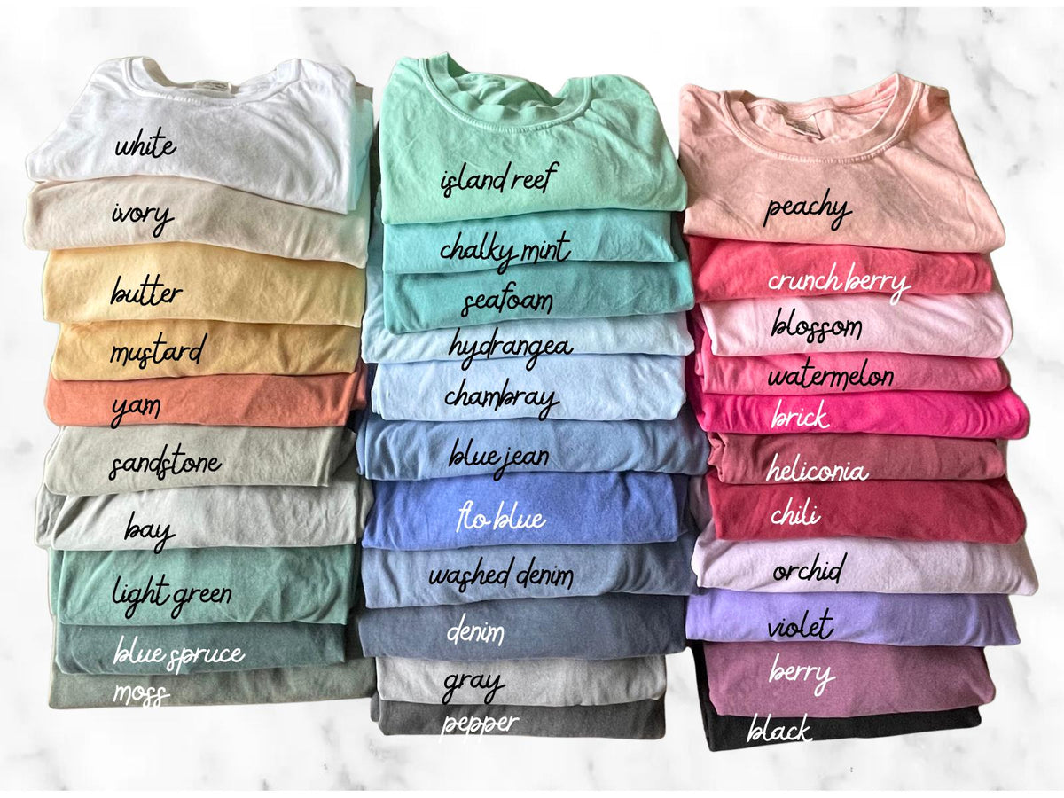 May include: A collection of folded crew neck t-shirts arranged in three vertical stacks, showcasing a wide variety of solid colors. Each shirt features a handwritten label identifying its specific color name, ranging from white to black.