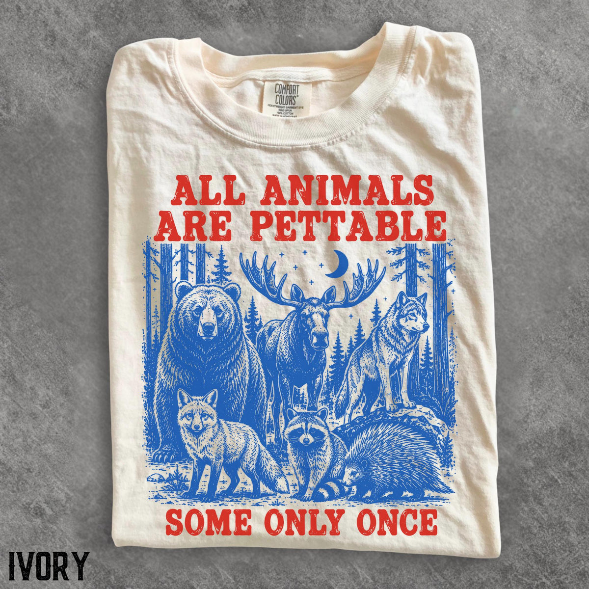 May include: An ivory t-shirt featuring a red graphic print that reads ALL ANIMALS ARE PETTABLE SOME ONLY ONCE. Below the text is a blue illustration of a bear, moose, wolf, fox, raccoon, and porcupine in a forest setting with a crescent moon.
