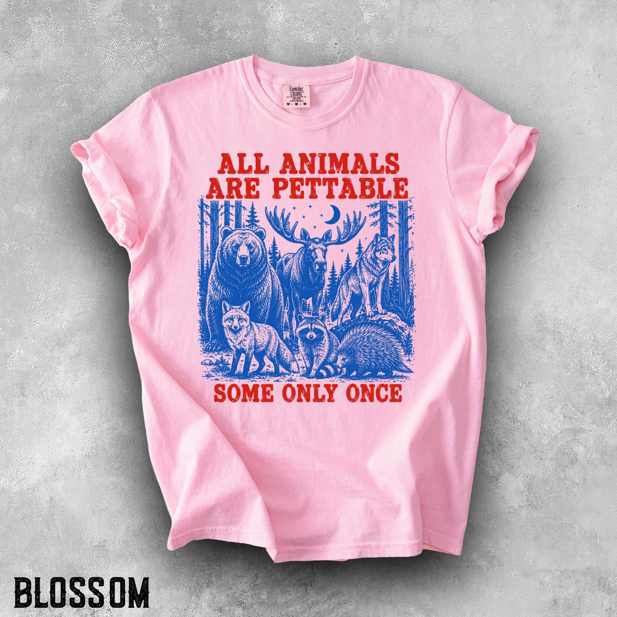 May include: A light pink short-sleeve t-shirt featuring a blue graphic of a bear, moose, wolf, fox, raccoon, and porcupine in a forest. The red text reads ALL ANIMALS ARE PETTABLE and SOME ONLY ONCE. The shirt has a crew neckline and short sleeves.
