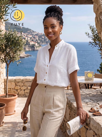 May include: A white short-sleeved button-down shirt with a mandarin collar and beige high-waisted trousers. The shirt features a front button placket and cuffed sleeves. The outfit is paired with gold hoop earrings and brown sunglasses.