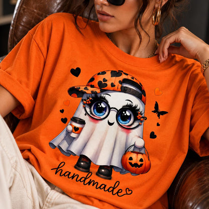May include: An orange t-shirt featuring a graphic of a white ghost wearing a baseball cap with black hearts, glasses, and long eyelashes. The ghost holds a coffee cup and a jack-o'-lantern bucket, surrounded by black hearts and a butterfly.