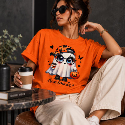 May include: An orange t-shirt featuring a graphic of a cartoon ghost wearing a baseball cap and glasses, holding a coffee cup and a pumpkin bucket. The design includes small black hearts and a butterfly, with the word handmade written in cursive script.