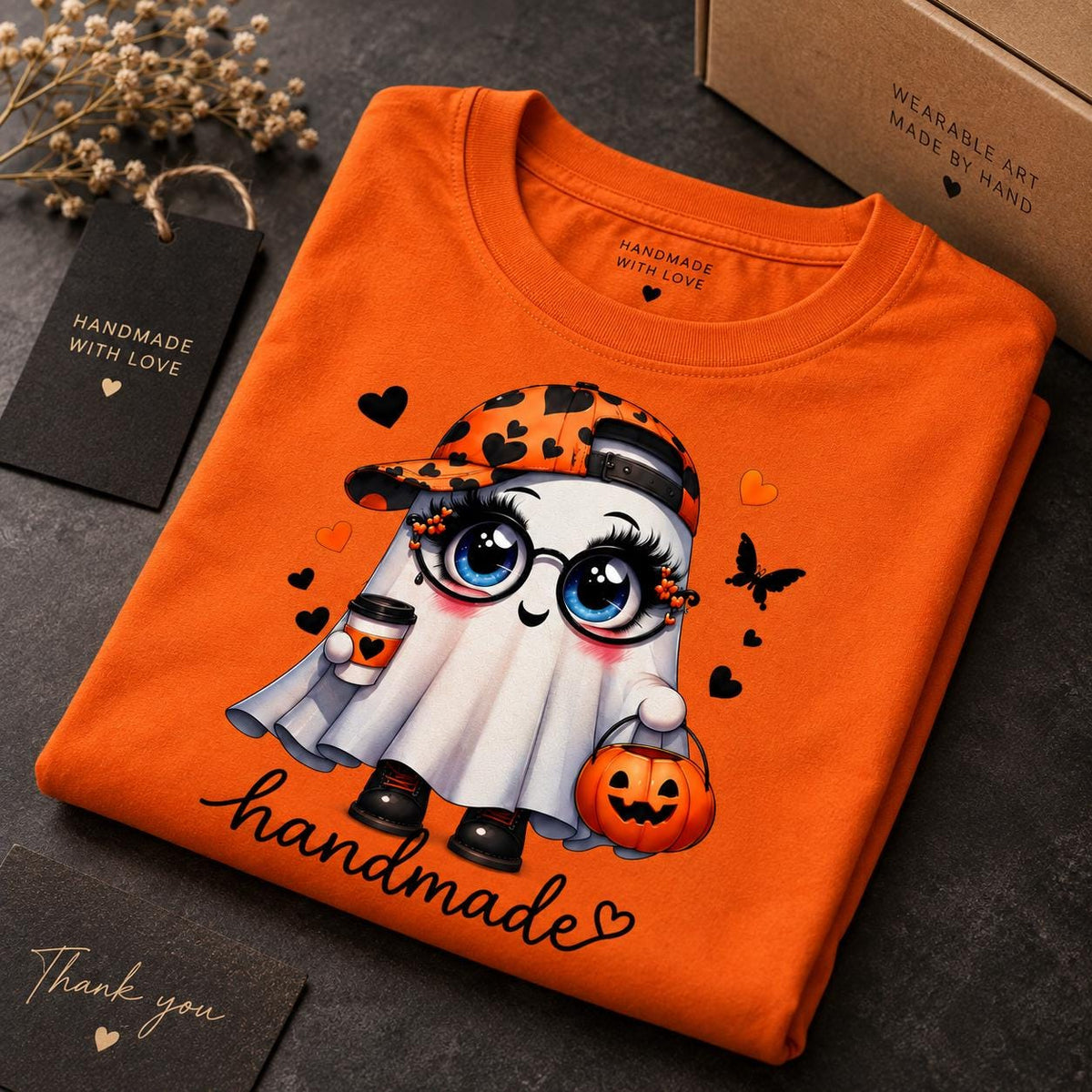 May include: An orange t-shirt featuring a cartoon ghost wearing a black and orange heart-patterned baseball cap and round glasses. The ghost holds a coffee cup and a jack-o'-lantern bucket. The word handmade is printed in black script below.