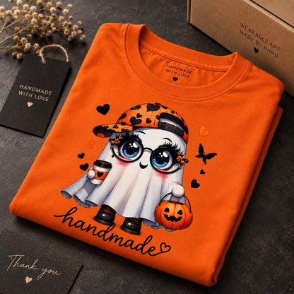 May include: An orange t-shirt featuring a cartoon ghost wearing a black and orange heart-patterned baseball cap and round glasses. The ghost holds a coffee cup and a jack-o'-lantern bucket. The word handmade is printed in black script below.
