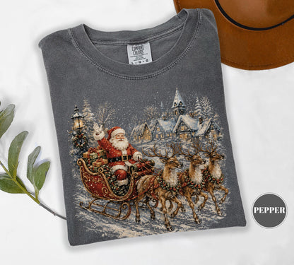 May include: A charcoal gray short-sleeve t-shirt featuring a graphic print of Santa Claus in a red sleigh pulled by three reindeer. The scene includes a snowy village with a church and a streetlamp. The shirt has a Comfort Colors brand label.