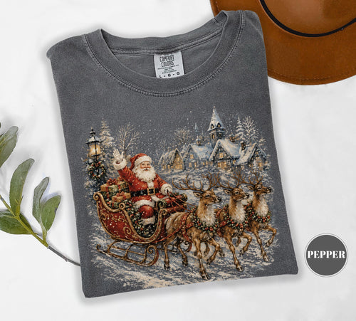 May include: A charcoal gray short-sleeve t-shirt featuring a graphic print of Santa Claus in a red sleigh pulled by three reindeer. The scene includes a snowy village with a church and a streetlamp. The shirt has a Comfort Colors brand label.