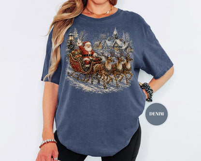May include: A denim blue short-sleeve t-shirt featuring a detailed graphic print of Santa Claus in a sleigh pulled by three reindeer. The scene includes a snowy village background with houses and trees. A small circular badge reads DENIM.