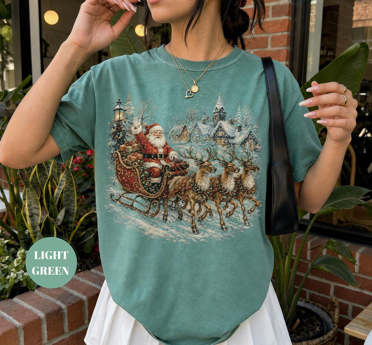 May include: A light green short-sleeve t-shirt featuring a detailed graphic print of Santa Claus in a sleigh pulled by three reindeer, traveling through a snowy village landscape with houses and trees under a winter sky.
