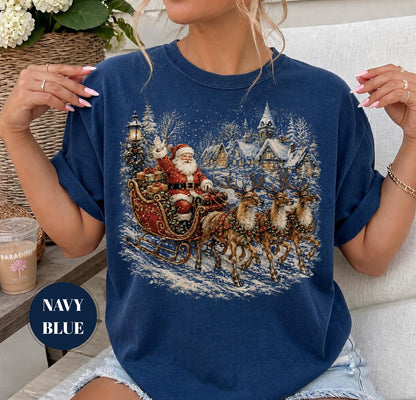 May include: A navy blue t-shirt featuring a graphic print of Santa Claus in a red sleigh pulled by three reindeer. The scene depicts a snowy village with houses and trees in the background. The design includes festive details and winter elements.
