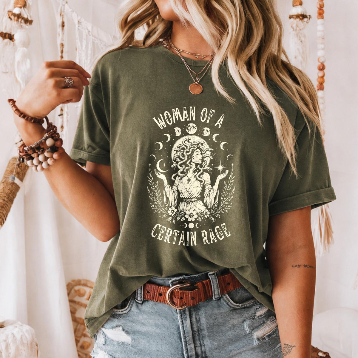 May include: An olive green short-sleeve t-shirt featuring a cream-colored graphic print of a figure with snake-like hair, surrounded by moon phases, stars, and foliage. The text reads "WOMAN OF A CERTAIN RAGE" in a distressed, arched font.