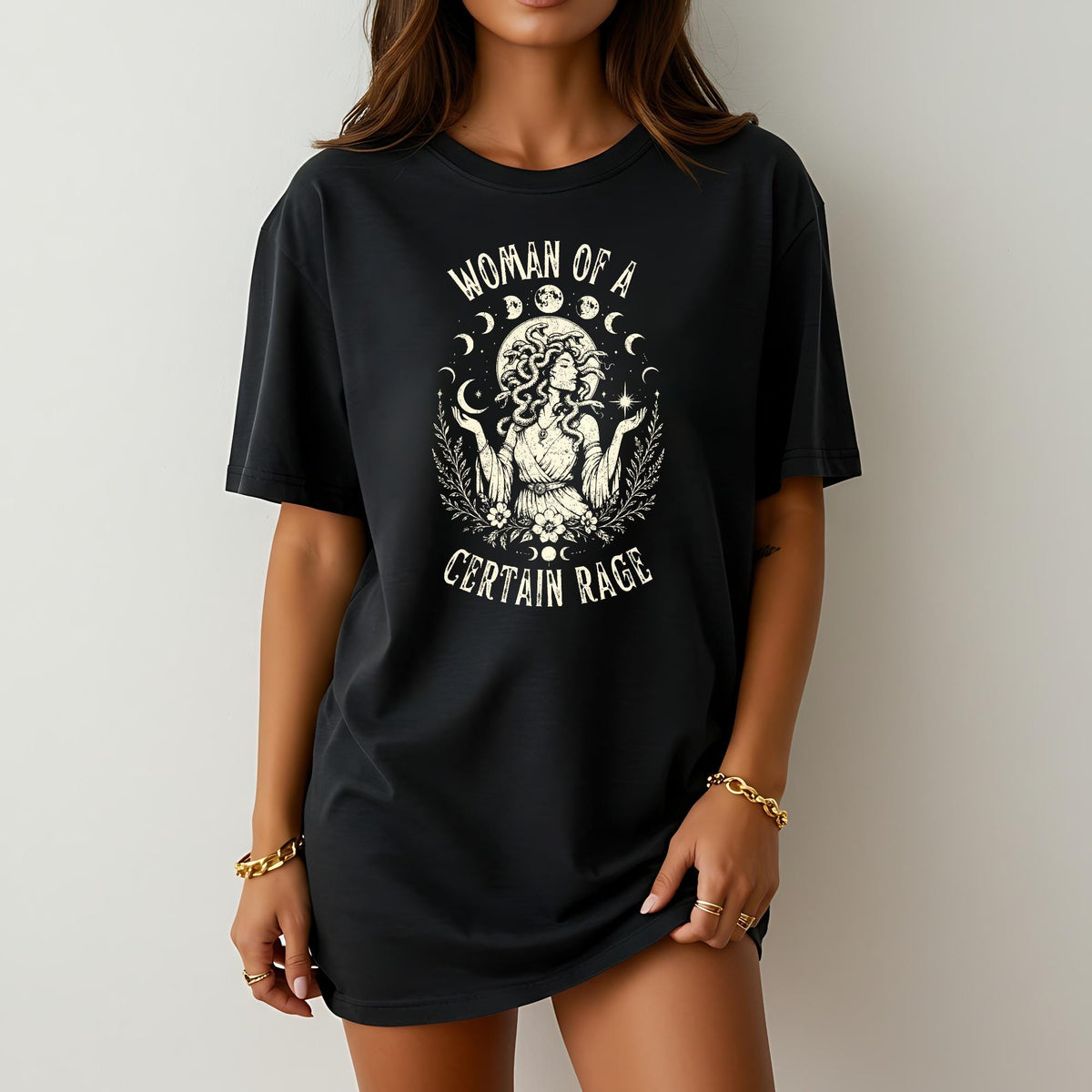 May include: A black short-sleeve t-shirt featuring a cream-colored graphic print of a figure with snakes for hair, surrounded by moon phases, stars, and foliage. The text "WOMAN OF A CERTAIN RAGE" is printed in a curved font above and below the figure.
