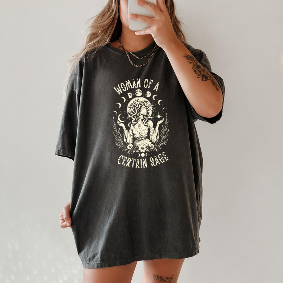 May include: A charcoal gray short-sleeve t-shirt featuring a cream-colored graphic print of a figure with snake-like hair surrounded by moon phases and floral elements, with the text "WOMAN OF A CERTAIN RAGE" arched above and below the illustration.