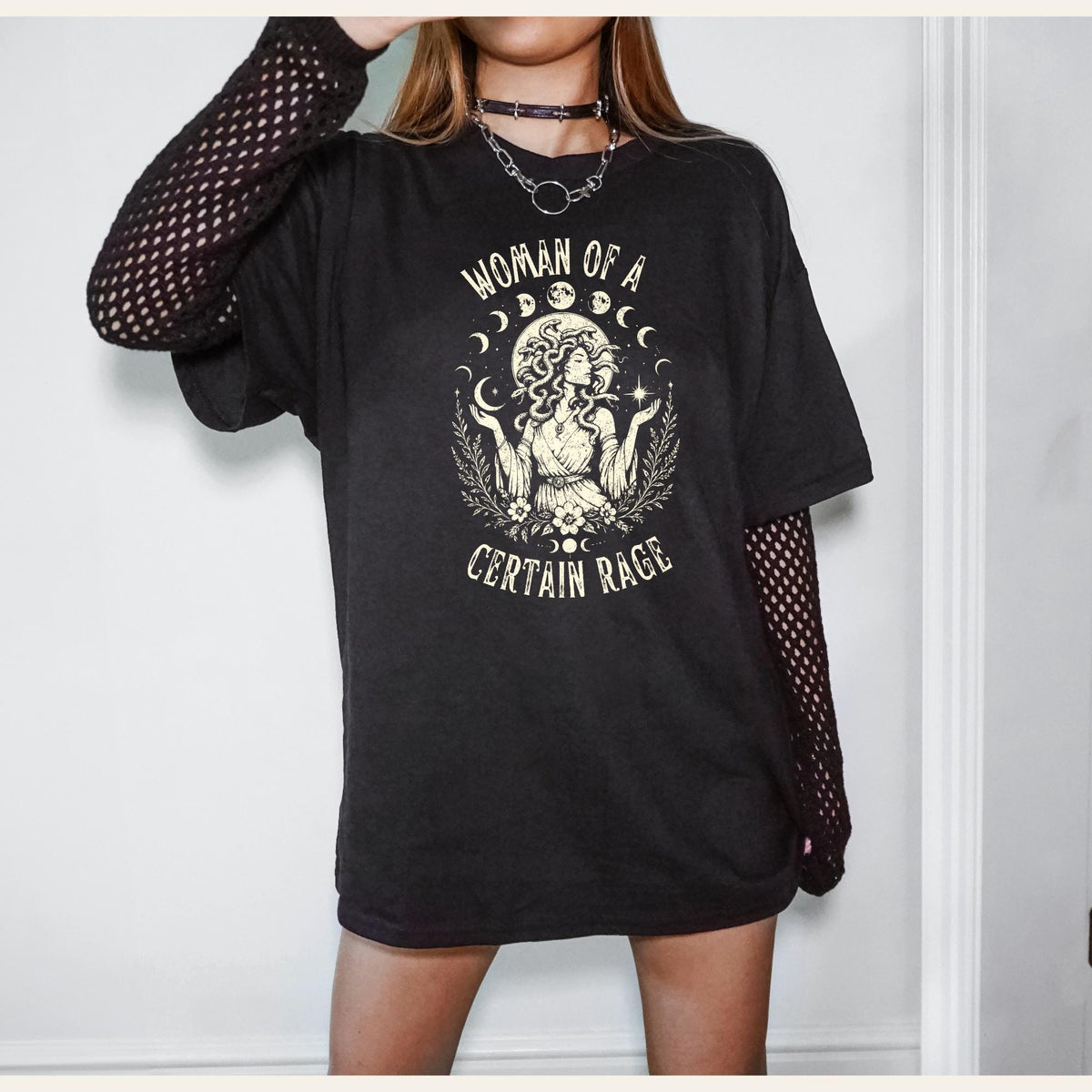 May include: A black short-sleeved t-shirt featuring a cream-colored graphic of a figure with snake-like hair, surrounded by moon phases and foliage, with the text "WOMAN OF A CERTAIN RAGE" printed in a curved arch above and below the illustration.
