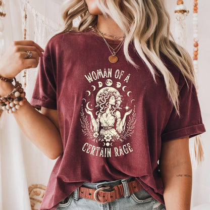 May include: A maroon short-sleeve t-shirt featuring a cream-colored graphic of a figure with snake-like hair, surrounded by moon phases and floral elements. The text "WOMAN OF A CERTAIN RAGE" is printed in a distressed font above and below the illustration.