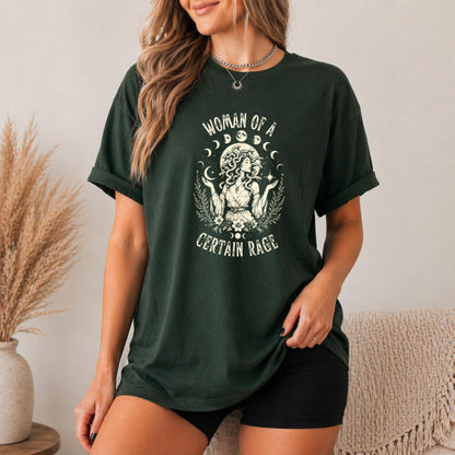 May include: A dark green short-sleeve t-shirt featuring a cream-colored graphic of a figure with snake-like hair surrounded by moon phases and foliage. The text "WOMAN OF A CERTAIN RAGE" is printed in a curved font above and below the central illustration.
