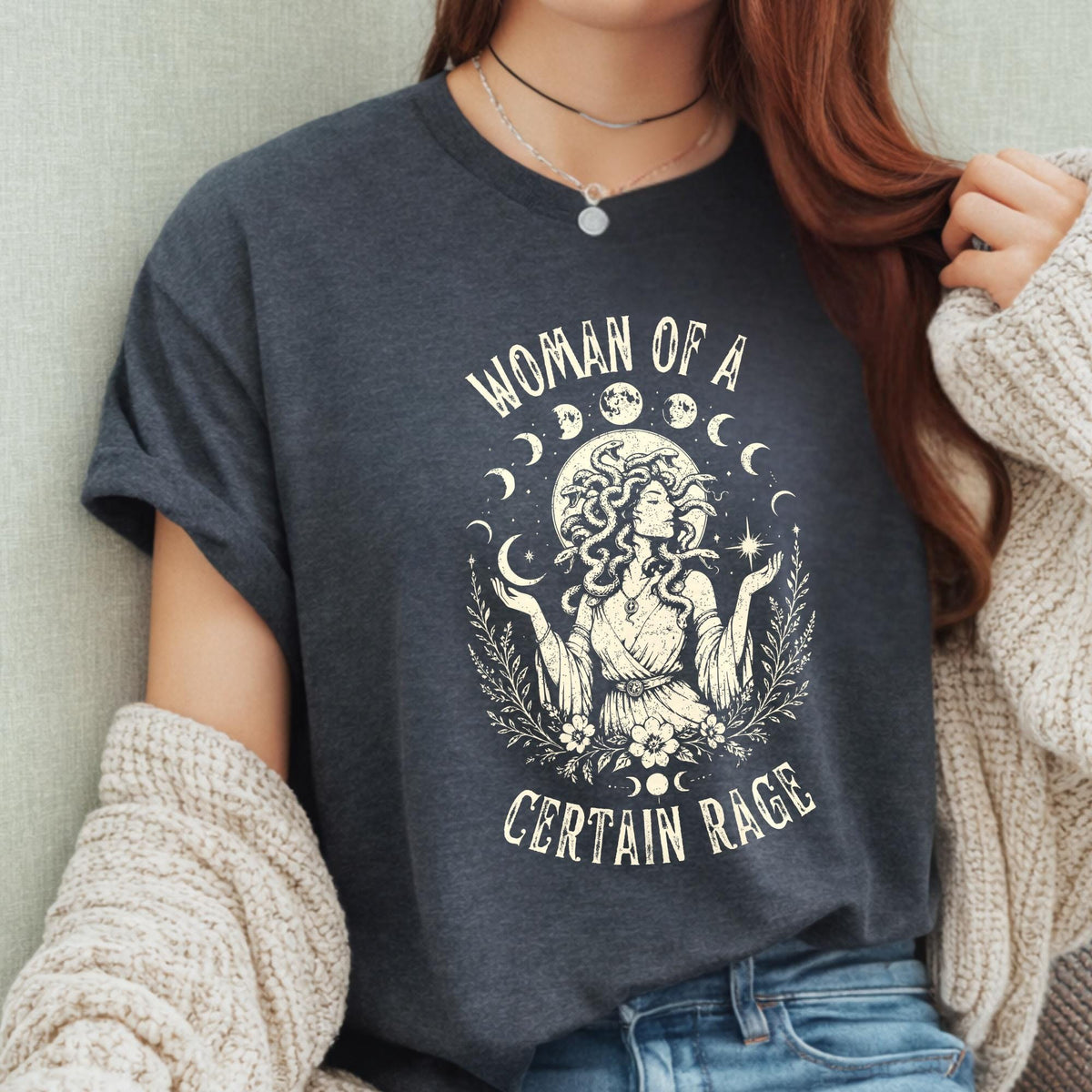 May include: A dark charcoal gray t-shirt featuring a cream-colored graphic print of a figure with snakes for hair, surrounded by moon phases, stars, and foliage. The text reads "WOMAN OF A CERTAIN RAGE" in a stylized font above and below the illustration.