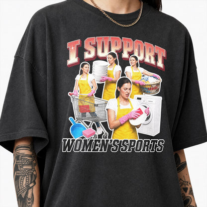 May include: A charcoal gray t-shirt featuring a graphic print with the text I SUPPORT WOMEN'S SPORTS. The design shows multiple figures wearing yellow aprons and pink gloves performing household chores like laundry, dishwashing, and cleaning.