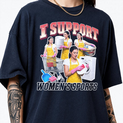May include: A dark navy blue t-shirt featuring a graphic print with the text I SUPPORT WOMEN'S SPORTS. The design shows a person in a yellow apron and pink gloves performing household chores like laundry, dishwashing, and shopping with a cart.
