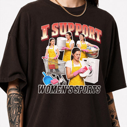 May include: A dark brown t-shirt featuring a graphic print with the text I SUPPORT WOMEN'S SPORTS. The design includes multiple images of a person wearing a yellow apron and pink gloves performing household chores like laundry and cleaning.