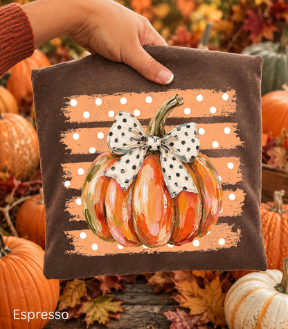 May include: A dark brown fabric square featuring a central graphic of an orange pumpkin with visible brushstroke textures. The pumpkin is adorned with a white bow featuring black polka dots, set against a background of horizontal orange stripes with white dots.