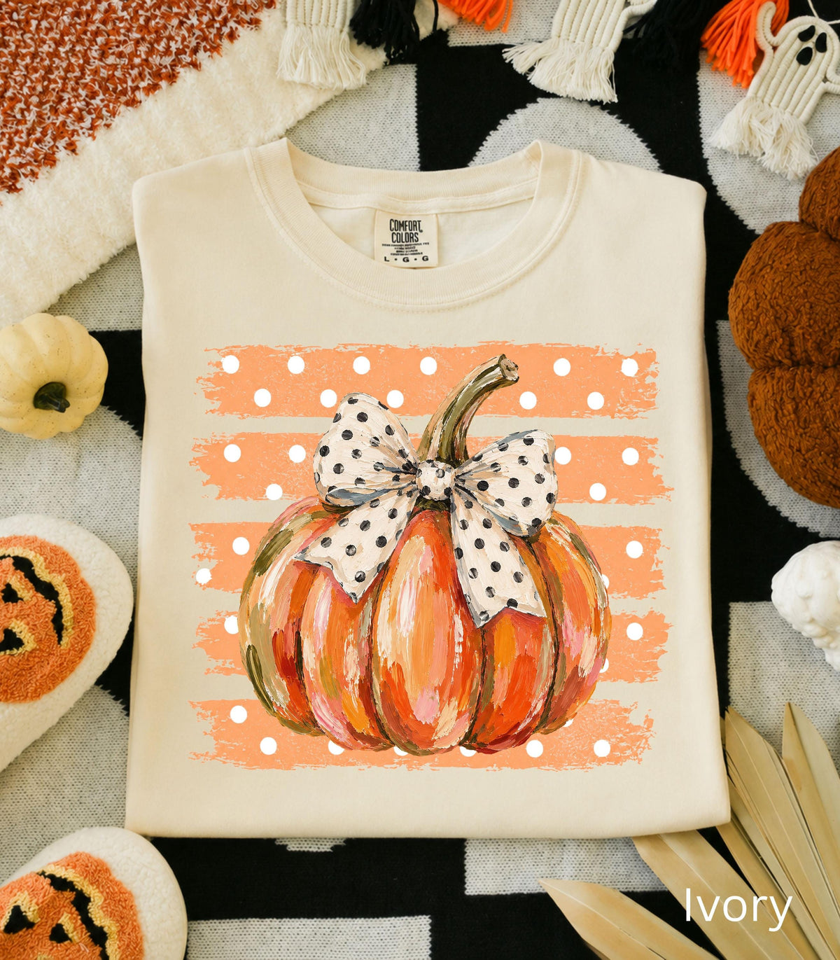 May include: An ivory crewneck t-shirt featuring a graphic of an orange pumpkin with a white and black polka dot bow. The pumpkin is centered over a background of horizontal orange brushstrokes with white polka dots. The shirt has a Comfort Colors label.