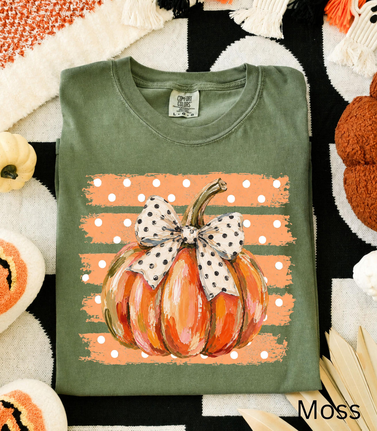 May include: An olive green t-shirt featuring a graphic of an orange pumpkin with a white and black polka dot bow. The pumpkin is centered over a rectangular background of horizontal orange brush strokes with white polka dots throughout the design.