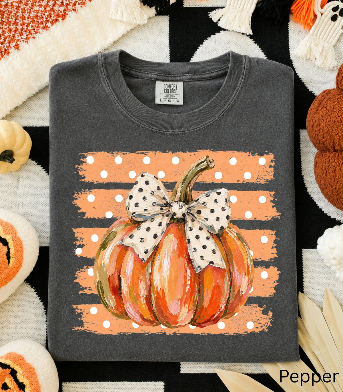 May include: A charcoal gray t-shirt featuring a graphic of an orange pumpkin with a white and black polka dot bow. The pumpkin is centered over a horizontal orange brushstroke background with white polka dots. The shirt has a Comfort Colors label.
