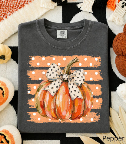 May include: A charcoal gray t-shirt featuring a graphic of an orange pumpkin with a white and black polka dot bow. The pumpkin is centered over a horizontal orange brushstroke background with white polka dots. The shirt has a Comfort Colors label.