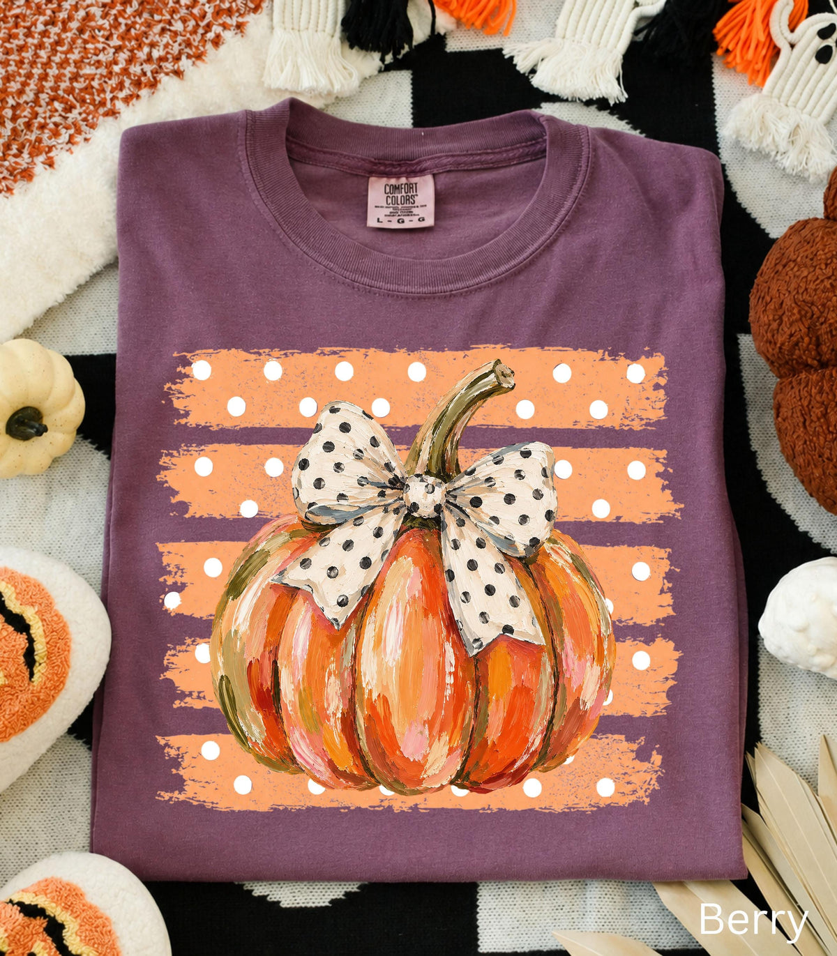 May include: A plum-colored t-shirt featuring a graphic of an orange pumpkin with a white and black polka-dot bow. The pumpkin is centered over a rectangular orange background with white polka dots. The shirt has a Comfort Colors brand label.
