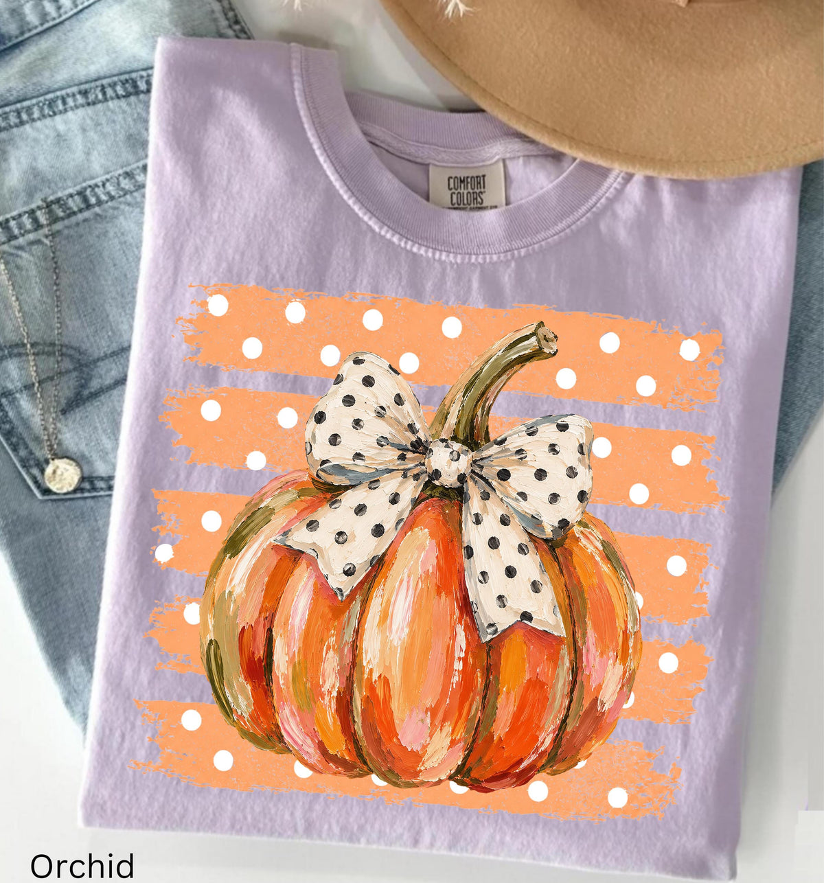 May include: A light purple t-shirt featuring a graphic of an orange pumpkin with a white and black polka dot bow. The pumpkin is centered over a background of horizontal orange brushstrokes with white polka dots. The shirt has a crew neckline.