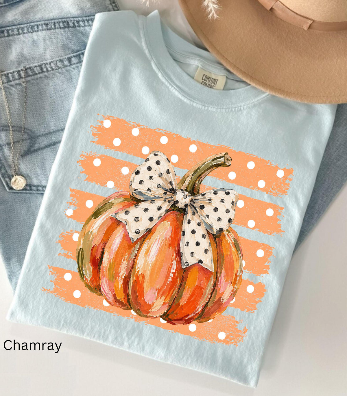 May include: A light blue t-shirt featuring a graphic of an orange pumpkin with a white and black polka dot bow. The pumpkin is centered over a background of horizontal orange brushstrokes with white polka dots. The shirt has a crew neckline.