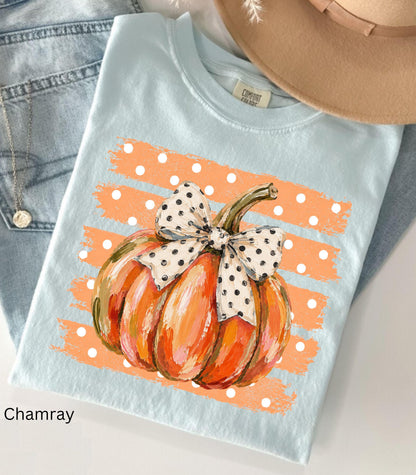 May include: A light blue t-shirt featuring a graphic of an orange pumpkin with a white and black polka dot bow. The pumpkin is centered over a background of horizontal orange brushstrokes with white polka dots. The shirt has a crew neckline.
