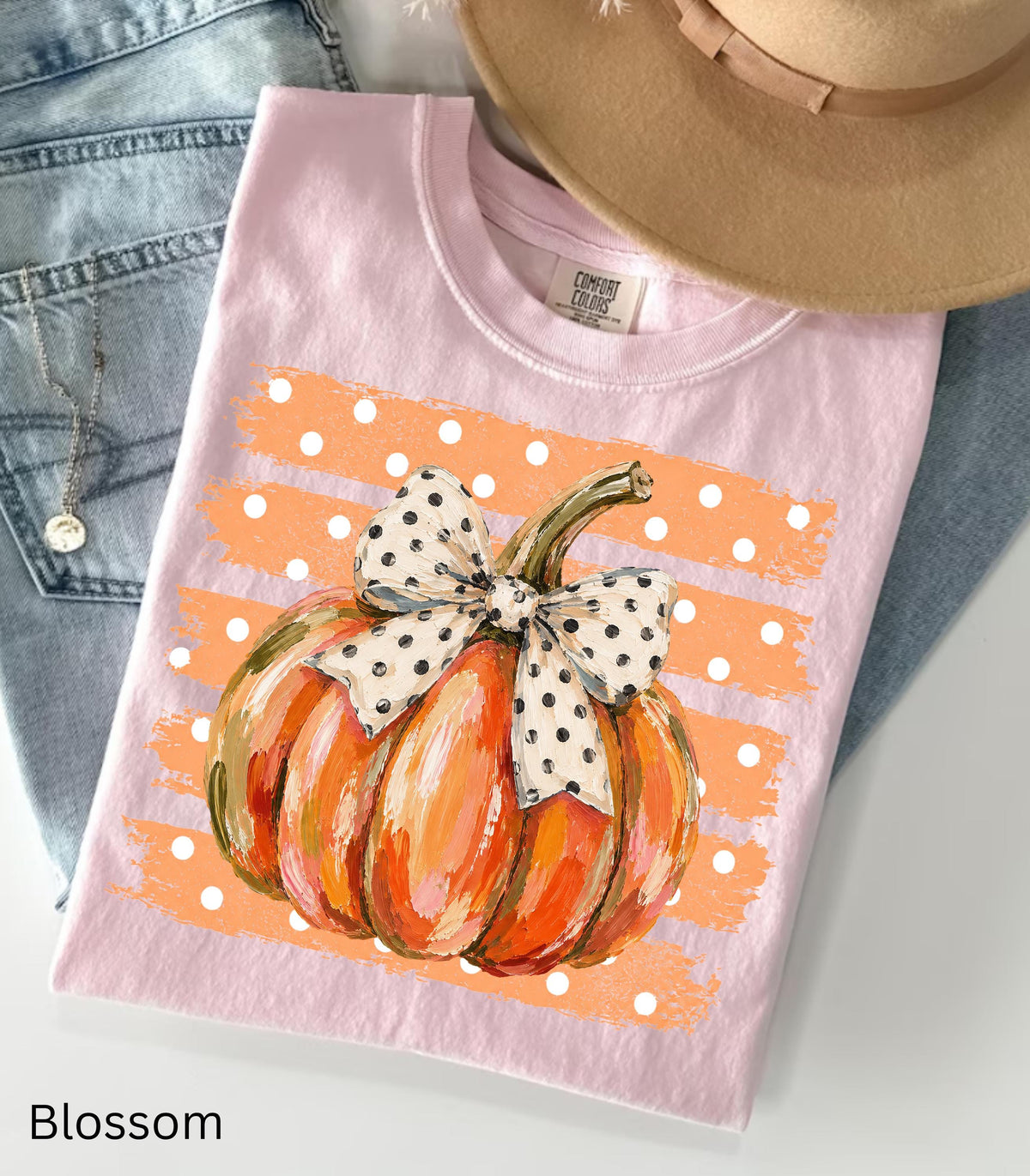 May include: A light pink t-shirt featuring a graphic of an orange pumpkin with a white and black polka dot bow. The pumpkin is centered over a horizontal orange brushstroke background with white polka dots. The shirt has a Comfort Colors brand label.