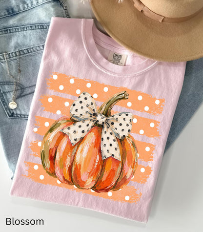 May include: A light pink t-shirt featuring a graphic of an orange pumpkin with a white and black polka dot bow. The pumpkin is centered over a horizontal orange brushstroke background with white polka dots. The shirt has a Comfort Colors brand label.