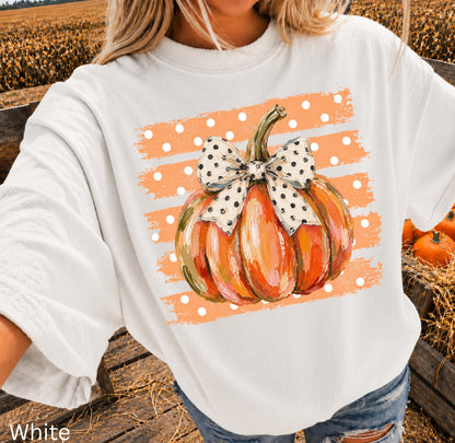 May include: A white short-sleeve t-shirt featuring a graphic of an orange pumpkin with a large white bow with black polka dots. The pumpkin is centered over a background of horizontal orange brush strokes with white polka dots throughout the design.