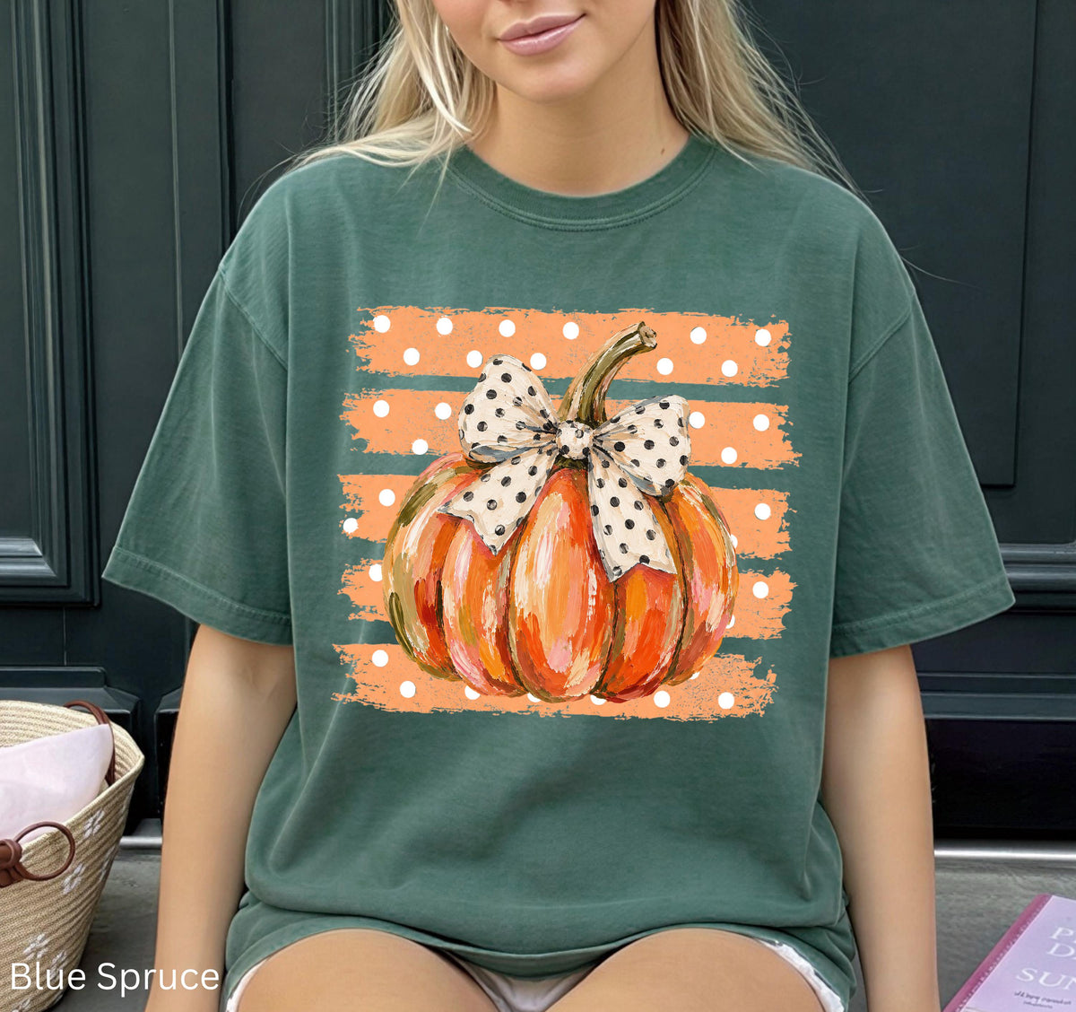 May include: A dark green short-sleeve t-shirt featuring a central graphic of an orange pumpkin with a white and black polka-dot bow. The pumpkin is set against a background of horizontal orange brushstroke stripes with small white polka dots.