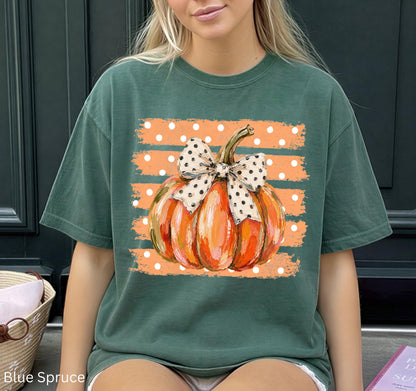 May include: A dark green short-sleeve t-shirt featuring a central graphic of an orange pumpkin with a white and black polka-dot bow. The pumpkin is set against a background of horizontal orange brushstroke stripes with small white polka dots.