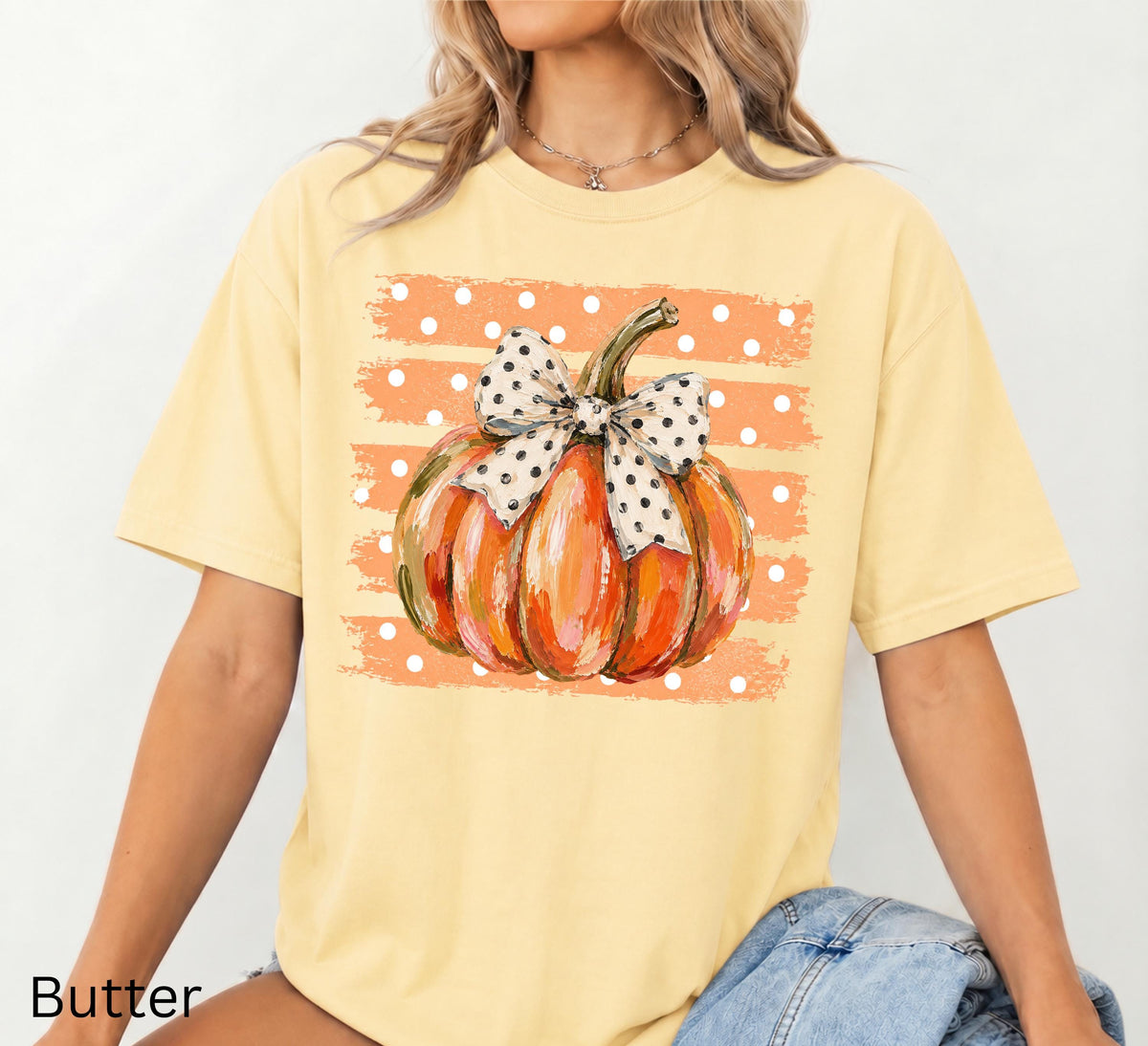 May include: A butter yellow short-sleeve t-shirt featuring a central graphic of an orange pumpkin with a white and black polka dot bow. The pumpkin is set against a background of horizontal orange brush strokes with white polka dots.
