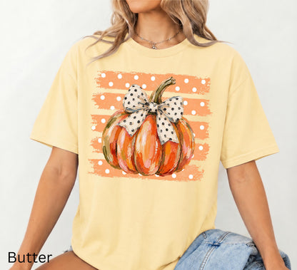 May include: A butter yellow short-sleeve t-shirt featuring a central graphic of an orange pumpkin with a white and black polka dot bow. The pumpkin is set against a background of horizontal orange brush strokes with white polka dots.