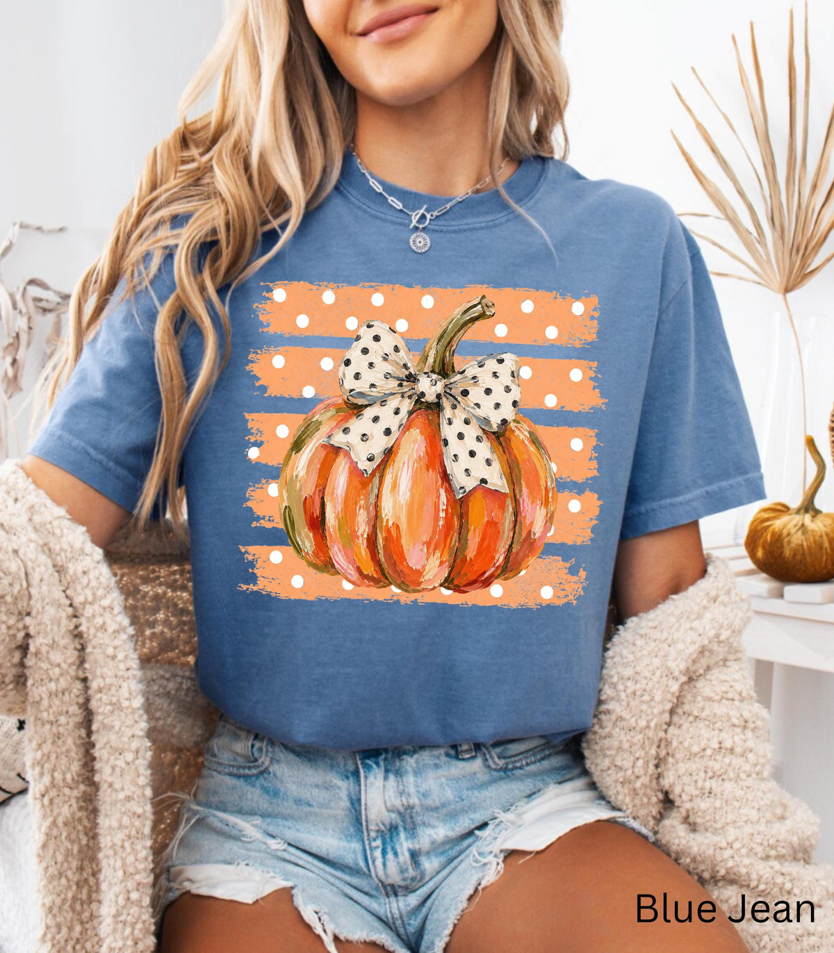 May include: A blue t-shirt featuring a graphic of an orange pumpkin with a white and black polka dot bow. The pumpkin is centered over a background of horizontal orange brush strokes with white polka dots. The text Blue Jean is printed below.