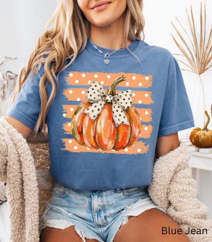 May include: A blue t-shirt featuring a graphic of an orange pumpkin with a white and black polka dot bow. The pumpkin is centered over a background of horizontal orange brush strokes with white polka dots. The text Blue Jean is printed below.