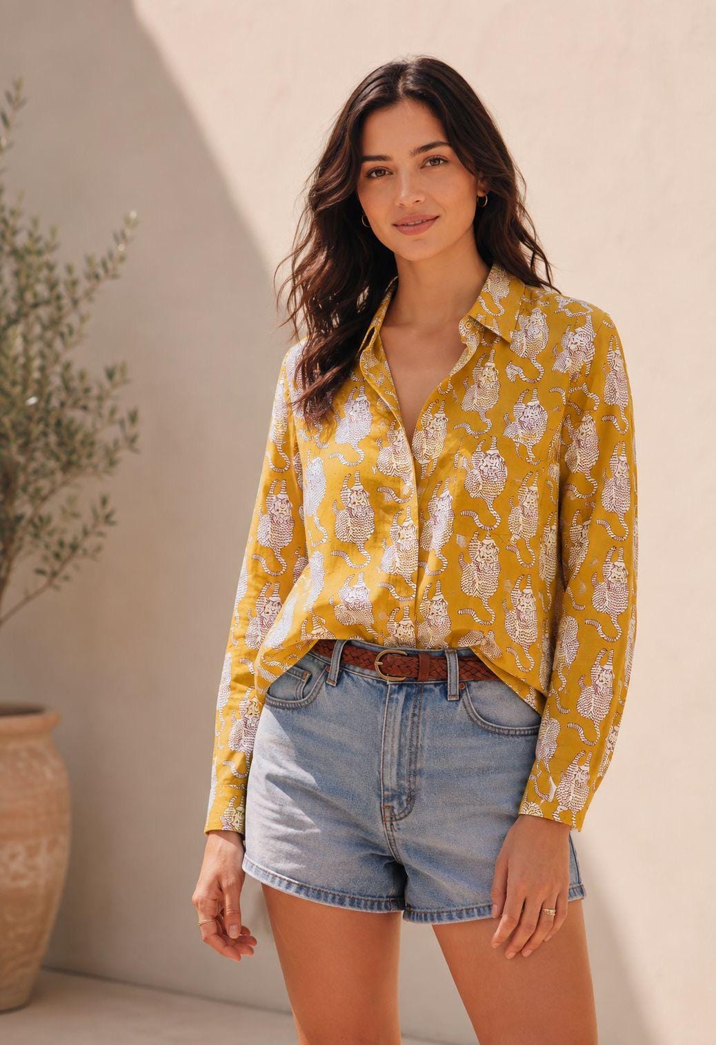 May include: A long-sleeved button-down shirt in mustard yellow featuring a repeating white and grey tiger print pattern, paired with light blue denim shorts and a brown woven leather belt with a gold-toned buckle.