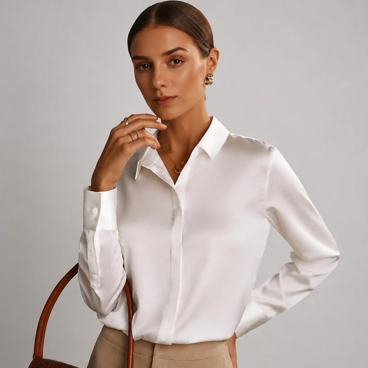 May include: A long-sleeved white button-down shirt made of a smooth, satin-like fabric with a classic collar, a front button placket, and structured cuffs. The shirt is paired with tan trousers and a brown leather handbag with a curved handle.