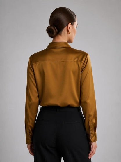 May include: A long-sleeved button-down shirt in a lustrous mustard gold satin fabric, featuring a classic collar, a back yoke, and buttoned cuffs, paired with solid black trousers. The shirt has a smooth, reflective finish and a tailored fit.